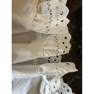 May include: Close-up of white fabric with eyelet lace trim. The fabric has a ruffled edge and a decorative cut-out pattern along the border. The image shows multiple layers of the fabric, creating a textured effect.