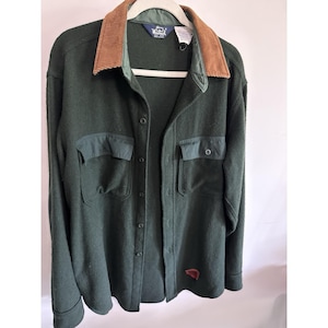 May include: A dark green button-up shirt with a corduroy collar and two chest pockets. The shirt has a small red and white logo on the lower right side. The collar is brown and the pockets are a darker green.