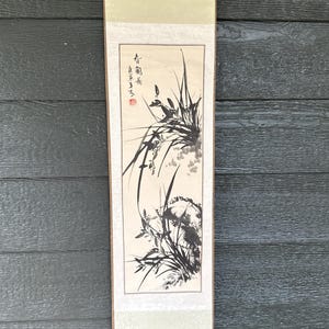 May include: A vertical hanging scroll featuring a black ink wash painting of orchids and rocks on a light beige paper background. The artwork includes black calligraphic text and a small red seal stamp. The scroll has cream-colored fabric borders.