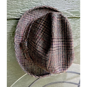 May include: A brown and black plaid hat with a rounded crown and a short brim. The hat is made of a textured fabric and has a classic, vintage style. The hat is resting on a metal surface.