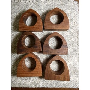 May include: A set of six brown wooden napkin rings, each featuring a pointed arch shape with a circular cutout in the center. The rings are arranged in two columns of three, displaying natural wood grain patterns and smooth, finished surfaces.