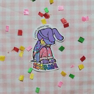 May include: A holographic sticker featuring a purple bunny wearing a blue hat and holding a sign that says "Miniature Mage Employment!" The sticker is on a pink and white checkered background.