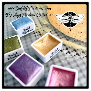 May include: Five watercolor paint pans in white square containers with the words "Sinful Reflections" on each. The pans are filled with metallic paints in shades of blue, green, purple, gold, and pink. The background is a textured surface with a dragonfly sticker.