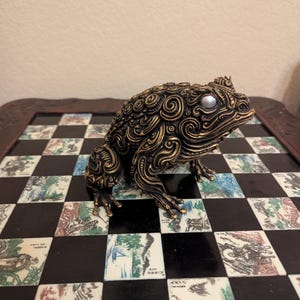 May include: A detailed bronze-colored frog figurine with gold accents and a pearl-like eye. The frog is intricately designed with swirling patterns and sits on a decorative chessboard with colorful squares.