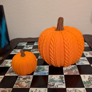 May include: Two orange pumpkins with a textured cable knit pattern and brown stems. The pumpkins are round and feature vertical ridges that mimic the appearance of knitted fabric, with one pumpkin significantly larger than the other.