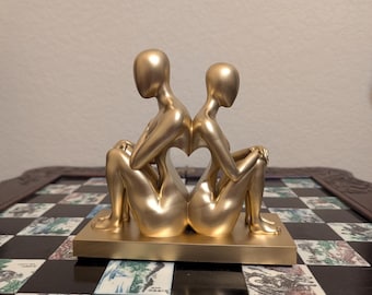 Puede incluir: Una escultura dorada de dos figuras sentadas de espaldas, formando un espacio en forma de corazón entre ellas. Las figuras están sobre una base rectangular. La escultura está colocada sobre un tablero de ajedrez decorativo.