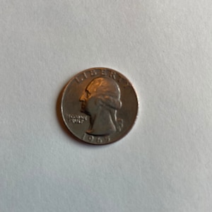 Op de afbeelding: Een zilveren Amerikaanse kwart dollar munt met het jaar 1965 gegraveerd aan de onderkant. De munt toont een profiel van George Washington en de woorden "LIBERTY" en "IN GOD WE TRUST" gegraveerd op de munt.