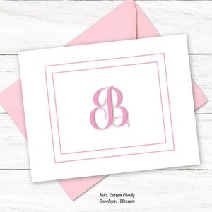 May include: A white stationery card with a pink double border and a pink letter "B" in the center, resting on a pink envelope. A silver pen with gold accents is to the right. The text "Ink: Cotton Candy, Envelope: Blossom" is at the bottom.