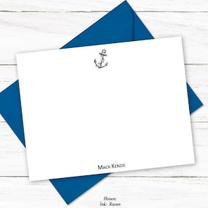 May include: A white stationery card with a small anchor graphic and the name "MACK KENZIE" printed at the bottom. The card is shown with a blue envelope and a silver pen on a white wood background.