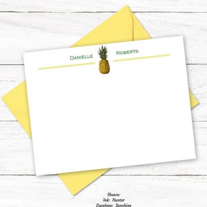 May include: White stationery with a pineapple illustration and the names "Danielle Roberts" in green. A yellow envelope and a silver pen with gold accents are also present. The stationery is from Big Dog Paper Co.