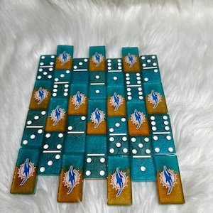 May include: A set of custom dominoes featuring a teal and gold color scheme. Each domino has white dots and a Miami Dolphins logo. The dominoes are arranged on a white, fluffy surface.