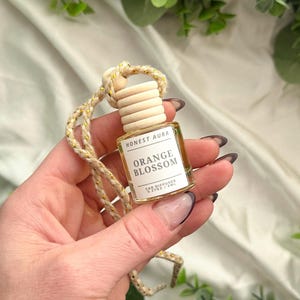 May include: A small glass bottle with a wooden cap and a gold string hanging from it. The bottle is filled with a clear liquid and has a label that reads "Honest Aura Orange Blossom Car Diffuser 0.27 oz / 8 mL".