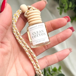 May include: A white car air freshener with a wooden cap and a gold and white string. The label reads "Honest Aura Clean Cotton Car Diffuser 0.27 oz / 8 ml."