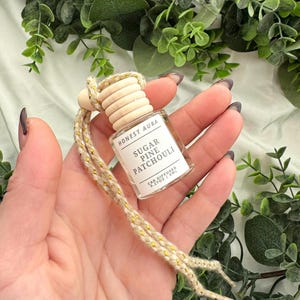 May include: A small glass bottle with a wooden cap and a gold and beige string. The bottle is labeled "Honest Aura Sugar Pine Patchouli Car Diffuser 0.2 fl oz / 7 ml".