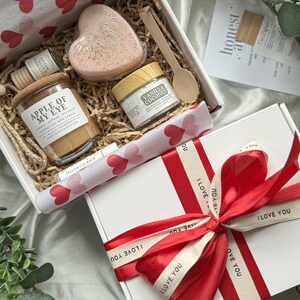 Könnte beinhalten: Eine weiße Geschenkbox mit einem roten Band und dem Aufdruck "Ich liebe dich". Die Box enthält eine kleinere weiße Box mit einer herzförmigen Seife, ein kleines Glas Vanille-Kokos-Körperbutter, einen Holzlöffel und eine Kerze mit dem Etikett "Apple of My Eye".