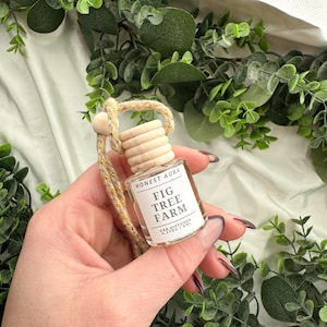 May include: A small glass bottle with a wooden cap and a gold string hanging from it. The bottle is labeled "Honest Aura Fig Tree Farm Car Diffuser 0.21 oz / 6 ml".