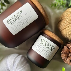May include: Two brown glass candles with cork lids, labeled "Sweater Season." The labels list scents like Cashmere, Sage, and Almond Milk. One candle is 10 oz, the other is 1 oz. The background includes greenery and a white pumpkin.