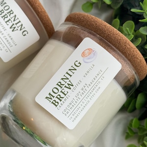 May include: A clear glass candle jar with a cork lid. The candle is white and has a label that reads "Morning Brew Caramel Coffee Vanilla Coconut Soy Wax Phthalate Free Fragrance 10 oz/283.4 g Sacramento, CA HONESTAURACO.ETSY.COM".