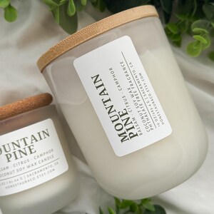 May include: Two frosted glass candle jars with wooden lids. The labels read "Mountain Pine" and include the scent notes "balsam, citrus, camphor" and "coconut soy wax candle".