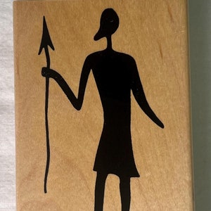 May include: A wooden block featuring a black silhouette of a figure holding a spear. The figure has a long neck and is wearing a short skirt. The background is a light brown color.