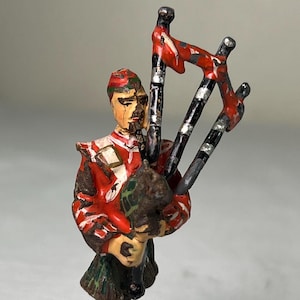 May include: A painted tin toy soldier figurine of a piper wearing a red and green kilt and holding a bagpipe with red, black, and white details.