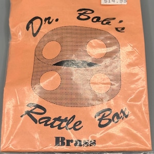 May include: An orange, sealed package featuring the text "Dr. Bob's Rattle Box Brass" in black script. The design includes a square shape with four circles and a central oval. The package is sealed in clear plastic.
