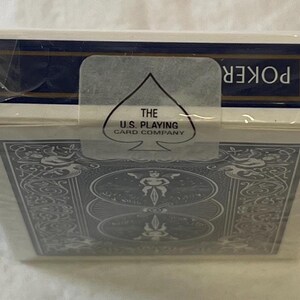 May include: Sealed deck of poker playing cards in a white box with a blue border. The box features an ornate design and the text "POKER". The U.S. Playing Card Company logo is visible on a clear seal.