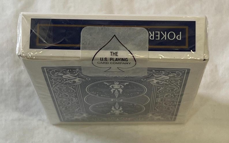 May include: Sealed deck of poker playing cards in a white box with a blue border. The box features an ornate design and the text "POKER". The U.S. Playing Card Company logo is visible on a clear seal.