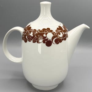 May include: White ceramic teapot with a brown floral design. The teapot has a curved handle, a long spout, and a lid. The floral pattern is on the upper portion of the teapot.