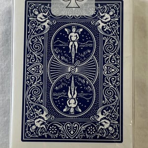 May include: A sealed deck of playing cards with a white border and a navy blue back design. The back features a central illustration of a person riding a bicycle, surrounded by ornate scrollwork and cherubs. The text "U.S. Playing Card Company" is visible.