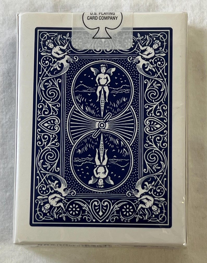 May include: A sealed deck of playing cards with a white border and a navy blue back design. The back features a central illustration of a person riding a bicycle, surrounded by ornate scrollwork and cherubs. The text "U.S. Playing Card Company" is visible.
