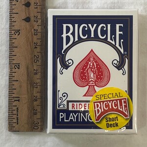 May include: A Bicycle playing card deck with a blue and white design. The deck features a red spade symbol with a figure in the center. A yellow sticker reads "Special Bicycle Short Deck". A wooden ruler is next to the deck.