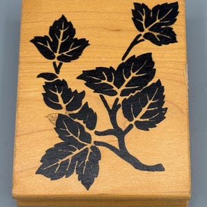 May include: A rectangular wooden stamp featuring a black leaf and branch design. The leaves have detailed veining, and the stamp has a light brown color. The design is centered on the stamp.