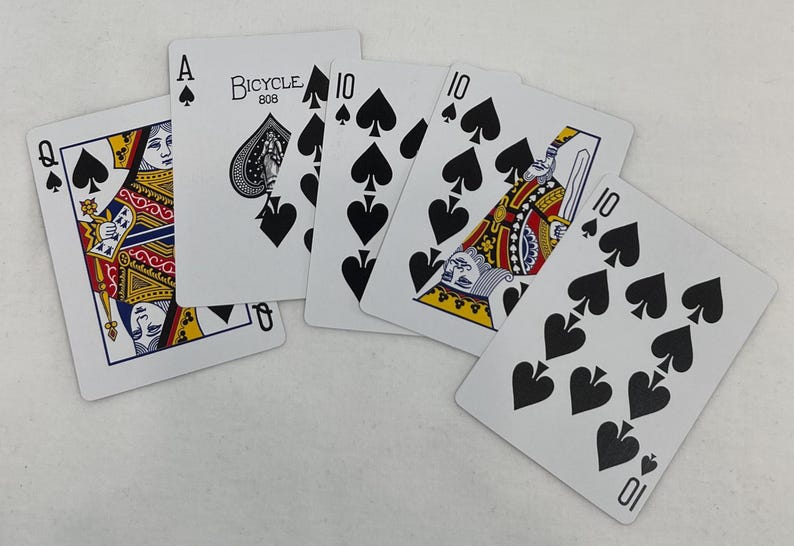 May include: A spread of playing cards featuring the Queen of Spades, Ace of Spades, and several 10 of Spades cards. The Bicycle brand is visible on the Ace of Spades. The cards are white with black spade symbols and colorful court card designs.