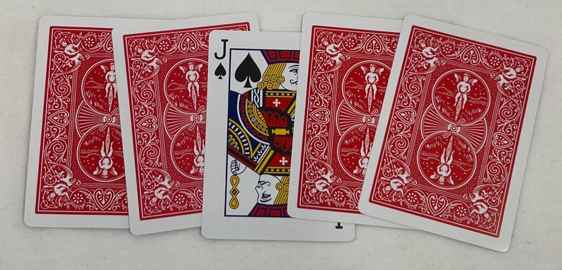 May include: A collection of playing cards featuring a Jack of Spades card in the center, flanked by four cards with red backs. The Jack of Spades card displays a detailed illustration with black, gold, red, and blue colors. The backs of the cards have a red ornate design.