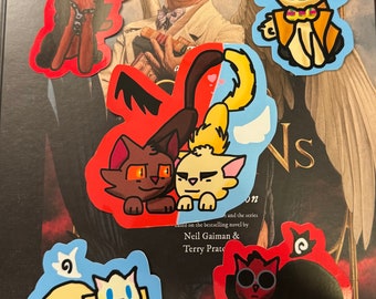 May include: A selection of cat-themed stickers in various colours. The stickers depict cartoon cats with different designs, such as angel wings, bow ties, and sunglasses. The stickers are placed on a book cover with the text "Good Omens".