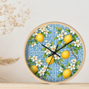 May include: Round wall clock with a light wood frame. The clock face features a blue and white tile design with yellow lemons, white flowers, and green leaves. Black clock hands show the time.