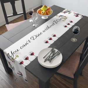 May include: White table runner with the text "You can't Dine without Wine" and illustrations of wine glasses, grapes, and wine bottles. The runner is on a dark wood table, set with plates, silverware, and wine glasses.