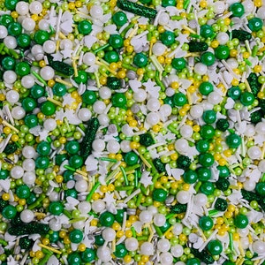 Könnte beinhalten: Nahaufnahme einer Mischung aus bunten Streuseln in Grün-, Weiß-, Gelb- und Silbertönen. Die Streusel umfassen kleine Kugeln, Sternformen und Stäbchenformen, wodurch ein festliches und strukturiertes Aussehen entsteht. Ideal zum Dekorieren von Backwaren.
