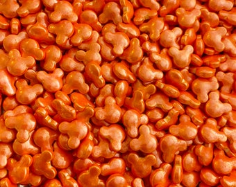 May include: Close-up of orange, Mickey Mouse-shaped sprinkles. The sprinkles are glossy and have a metallic sheen. The image is a full frame of the sprinkles, with no other objects visible. The sprinkles are a uniform colour and shape, creating a visually appealing texture.