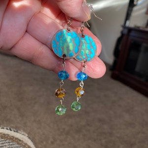 Op de afbeelding: Een paar oorhangers met ronde blauwe en groene gepatroneerde metalen schijven en hangende blauwe, groene en amberen kralen.