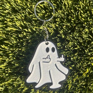 May include: A white ghost-shaped keychain with a black border, giving the middle finger. The keychain has a silver ring and chain. The ghost has two black eyes and a black outline of a hand giving the middle finger.