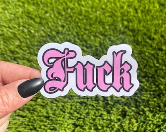 May include: A sticker with the word "Fuck" in a pink gothic font with a black outline. The sticker is on a white background and held against a green background.