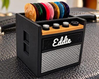 May include: A black guitar pick holder shaped like a miniature amplifier with the name Eddie in white script on the front. The top slot holds several colorful guitar picks in shades of orange, green, red, pink, brown, blue, and purple.