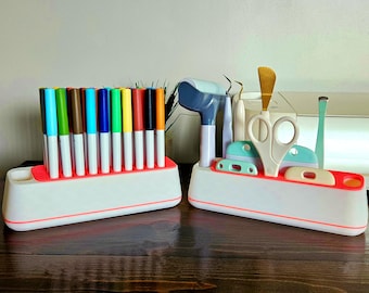 Puede incluir: Dos organizadores blancos con acentos rojos. Un organizador contiene una variedad de marcadores de colores, el otro contiene artículos de artesanía, incluyendo tijeras, un rodillo de pintura y una pequeña herramienta.