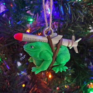 May include: A green frog Christmas ornament with a silver missile strapped to its back. The missile has the words "MISSILE TOAD" printed on it.
