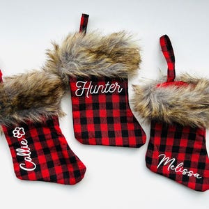 Puede incluir: Tres calcetines de Navidad de cuadros rojos y negros con ribete de piel sintética. Los calcetines están personalizados con los nombres "Hunter", "Callie" y "Melissa".