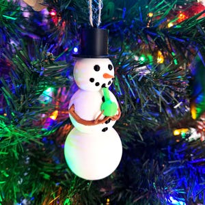 Puede incluir: Un adorno de Navidad de muñeco de nieve blanco con un sombrero de copa negro y una pipa de vidrio verde en la mano.