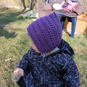 Könnte beinhalten: Ein Kind trägt eine lilafarbene Häkelmütze und eine schwarze Jacke mit Blumenmuster. Das Kind hält eine durchsichtige Plastiktasse.
