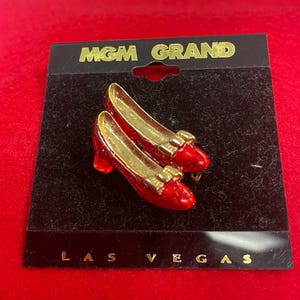 May include: A red and gold shoe-shaped pin with the text "MGM GRAND LAS VEGAS" on the black backing. The pin features two red shoes with gold heels and bows, likely a souvenir or collectible item.